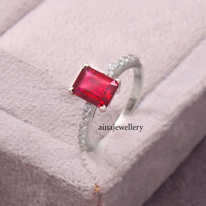 K&ouml;nnte beinhalten: Ein silberner Ring mit einem gro&szlig;en, rechteckigen, leuchtend roten Edelstein in der Mitte, flankiert von kleineren, klaren, funkelnden Steinen, die entlang der Ringschiene eingelassen sind. Der Text ainajewellery ist nahe der Basis sichtbar.