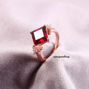 May include: Rose gold ring featuring a large, square-cut, deep red gemstone as the centerpiece. The ring is accented with smaller, clear gemstones on either side of the main stone. The ring is set against a soft, gray background.