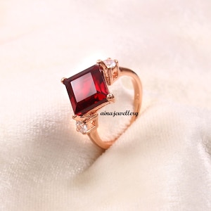 May include: Rose gold ring featuring a large, square-cut, deep red gemstone as the centerpiece. Two smaller, clear gemstones flank the main stone. The ring is set on a white surface, with the text "ainajewellery" visible.