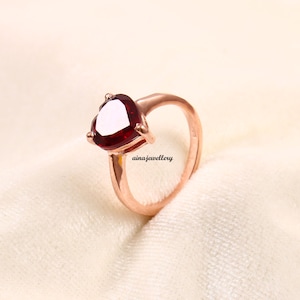 May include: A rose gold ring with a heart-shaped red gemstone. The ring is on a white background.
