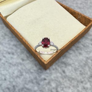 May include: A silver ring with a large oval-shaped red gemstone and two smaller white gemstones on either side.
