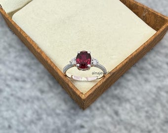 May include: A silver ring with a large oval-shaped red gemstone and two smaller white gemstones on either side.
