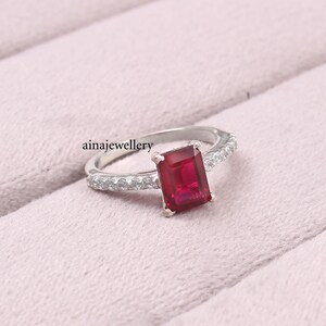 K&ouml;nnte beinhalten: Ein silberfarbener Ring mit einem rechteckigen, im Smaragdschliff geschliffenen roten Edelstein in der Mitte, der von vier Krappen gehalten wird. Die Ringschiene ist seitlich mit kleinen, runden, klaren Steinen besetzt. Text ainajewellery links.