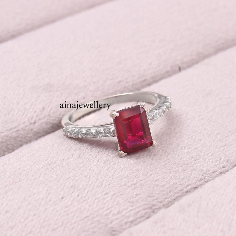 K&ouml;nnte beinhalten: Ein silberfarbener Ring mit einem rechteckigen, im Smaragdschliff geschliffenen roten Edelstein in der Mitte, der von vier Krappen gehalten wird. Die Ringschiene ist seitlich mit kleinen, runden, klaren Steinen besetzt. Text ainajewellery links.