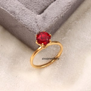 May include: A gold ring featuring a round, faceted, vibrant red gemstone held in a six-prong setting. The band is smooth and polished gold. The text "ainajewellery" is printed in black on the surface near the base of the ring.