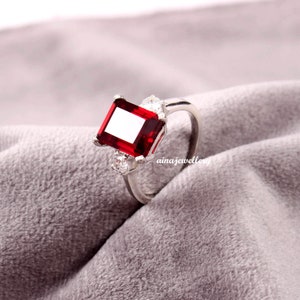 May include: A silver ring featuring a large, square-cut, deep red gemstone as the focal point. Two smaller, clear gemstones flank the main stone. The ring is set against a soft, gray fabric.