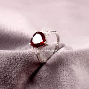 May include: A silver ring featuring a heart-shaped, deep red gemstone as the centerpiece. The ring is accented with smaller, clear gemstones on either side of the heart. The brand name "ainajewellery" is visible on the ring's band.