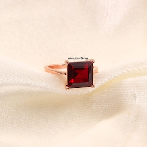 K&ouml;nnte beinhalten: Ein ros&eacute;goldener Ring mit einem gro&szlig;en, quadratischen, tiefroten Edelstein. Der Ring ist auf einem weichen, cremefarbenen Stoff platziert. Der Markenname "ainajewellery" ist sichtbar.