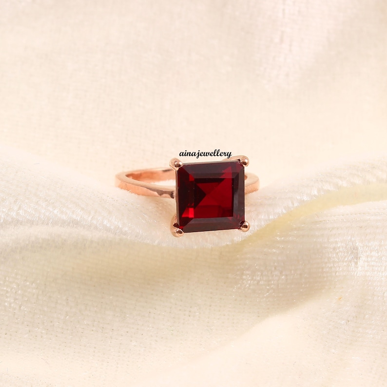 K&ouml;nnte beinhalten: Ein ros&eacute;goldener Ring mit einem gro&szlig;en, quadratischen, tiefroten Edelstein. Der Ring ist auf einem weichen, cremefarbenen Stoff platziert. Der Markenname "ainajewellery" ist sichtbar.