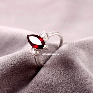 Könnte beinhalten: Ein silberner Ring mit einem roten, markisenförmigen Edelstein und drei kleineren, klaren Edelsteinen. Der Ring liegt auf einem grauen Stoffhintergrund.