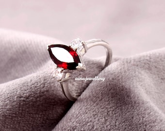 May include: A silver ring featuring a marquise-shaped red gemstone and three smaller clear gemstones. The ring is displayed on a grey fabric background.