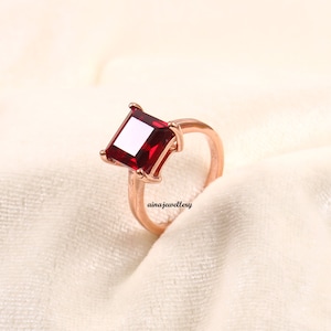 K&ouml;nnte beinhalten: Ein ros&eacute;goldener Ring mit einem gro&szlig;en, quadratisch geschliffenen, tiefroten Edelstein. Der Edelstein ist der Mittelpunkt, gefasst in einer Vier-Zacken-Fassung. Der Ring ist schlicht und poliert. Der Text "ainajewellery" ist sichtbar.