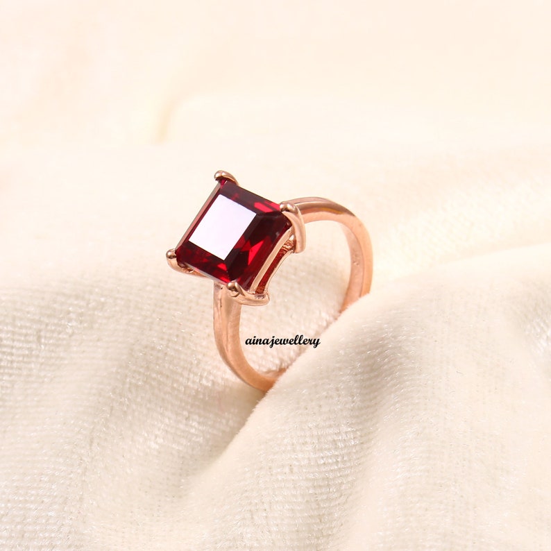 K&ouml;nnte beinhalten: Ein ros&eacute;goldener Ring mit einem gro&szlig;en, quadratisch geschliffenen, tiefroten Edelstein. Der Edelstein ist der Mittelpunkt, gefasst in einer Vier-Zacken-Fassung. Der Ring ist schlicht und poliert. Der Text "ainajewellery" ist sichtbar.