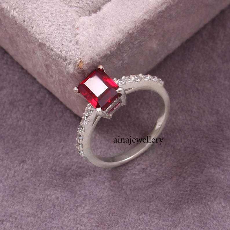 K&ouml;nnte beinhalten: Ein silberner Ring mit einem gro&szlig;en, rechteckigen, tiefroten Edelstein in der Mitte. Die Ringschiene ist auf beiden Seiten mit einer Reihe kleiner, klarer, funkelnder Steine besetzt. Der Text ainajewellery ist auf dem Ring zu sehen.