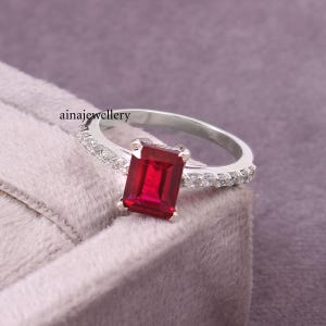 K&ouml;nnte beinhalten: Ein silberner Ring mit einem rechteckigen, im Smaragdschliff geschliffenen roten Edelstein als Mittelstein, flankiert von einer Reihe kleiner klarer Steine entlang der Ringschiene. Der Text ainajewellery ist auf der Innenseite des Rings sichtbar.