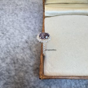 Może przedstawiać: Srebrny pierścionek z fioletowym kamieniem szlachetnym otoczonym halo mniejszych, przezroczystych kamieni szlachetnych. Pierścionek znajduje się w małym pudełku.