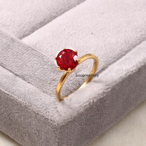 Könnte beinhalten: Ein Goldring mit einem runden, facettierten, leuchtend roten Edelstein in einer sechs-Krappen-Fassung. Die Ringschiene ist aus glattem, poliertem Gold. Der Text ainajewellery ist nahe der Basis der Fassung an der Seite des Rings sichtbar.