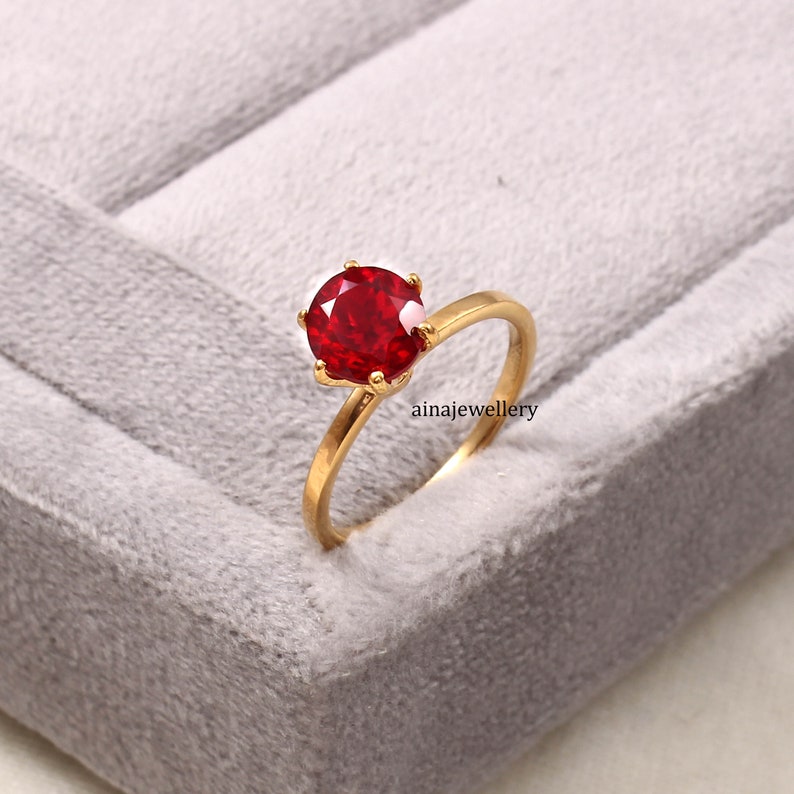 May include: A gold ring featuring a round, faceted, vibrant red gemstone held in a six-prong setting. The band is smooth and polished gold. The text ainajewellery is visible near the base of the ring setting on the side of the band.