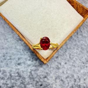 May include: A gold ring with an oval, deep red gemstone. The ring is set in a detailed, ornate band. The ring is displayed in a wooden box with a white interior. The text "ainajewelry" is visible on the ring.
