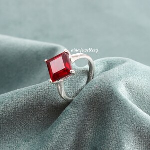 May include: A silver ring with a large, square, red gemstone. The ring is set on a blue velvet surface.