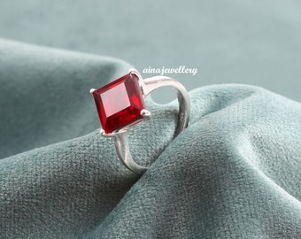 May include: A silver ring featuring a large, square, red gemstone. The ring is displayed on a blue velvet surface.