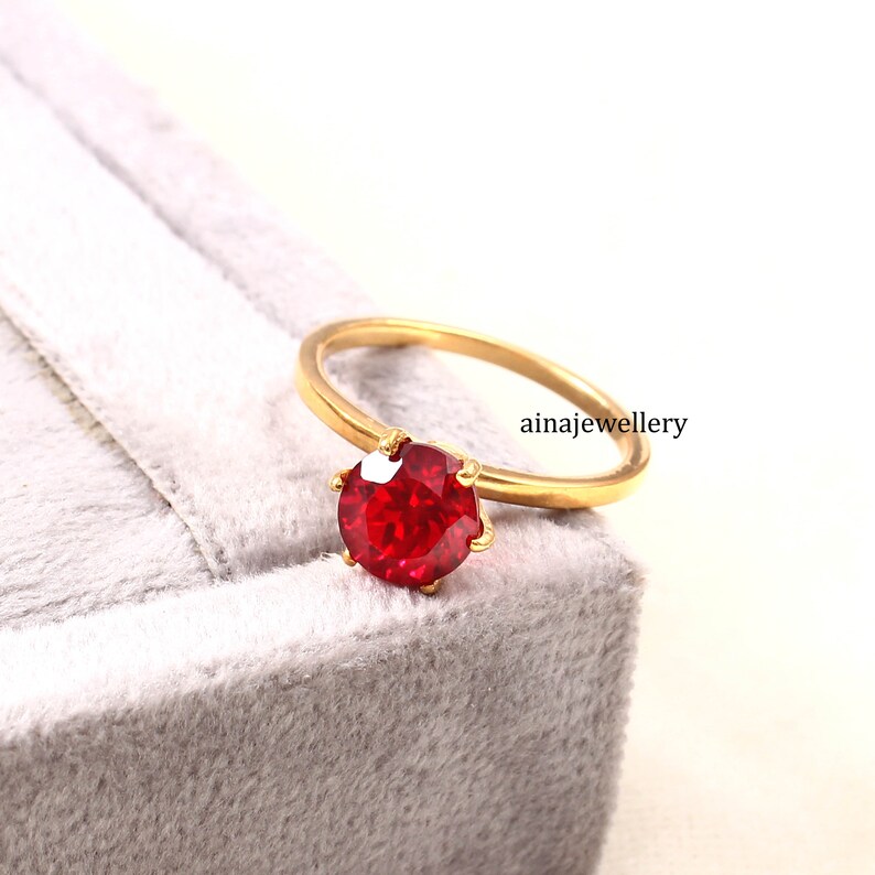 May include: A gold ring featuring a round, faceted, vibrant red gemstone held by six prongs. The band is smooth and polished gold. The text "ainajewellery" is visible near the ring, which is positioned on a soft, light grey textured surface.