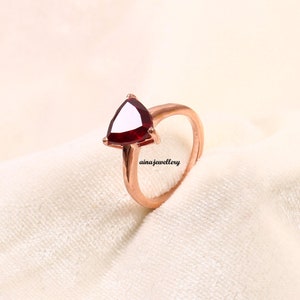 May include: A rose gold ring featuring a triangular, deep red gemstone. The ring's band curves gracefully, leading to the setting that holds the vibrant stone. The text "ainajewellery" is visible on the fabric.