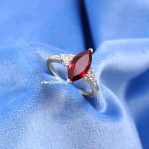 May include: A silver ring featuring a marquise-cut, deep red gemstone as the centerpiece. The ring is flanked by two triangular clusters of smaller, clear gemstones. The ring is set against a blue background. The text "ainajewellery" is visible on the ring.