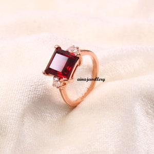 May include: A rose gold ring with a large, square, red gemstone and two smaller, round, white gemstones.