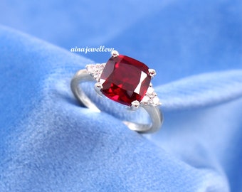 May include: A silver ring featuring a large, square-shaped, red gemstone surrounded by smaller, clear gemstones.