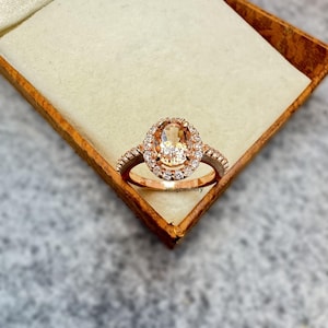 May include: Rose gold ring featuring an oval-cut peach-colored gemstone surrounded by a halo of small, clear stones. The band is also adorned with small stones. The ring is displayed in a wooden box.