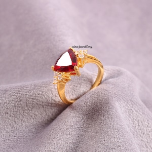 Puede incluir: Anillo de oro con una piedra roja triangular y pequeñas piedras transparentes. El anillo está sobre un fondo de tela gris.