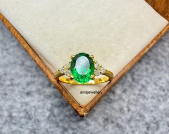 May include: A gold ring with a large, oval-cut emerald center stone and smaller diamond accents. The ring is set in a gold band and is displayed in a wooden jewelry box. The text "ainajewellery" is visible.