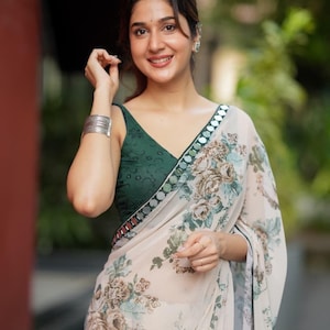 May include: A woman wearing a green top and a cream-colored sari with a floral pattern. The sari has a decorative border and is draped over the shoulder. The woman is smiling and wearing silver jewelry.