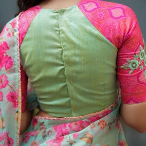 May include: A light green blouse with pink patterned sleeves and a matching floral print saree. The sleeves have a pink and orange geometric design with embroidered floral details. The saree features a light blue background with large pink flowers.