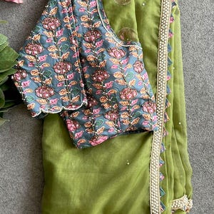 May include: A green saree with a floral patterned blouse. The blouse is a teal color with pink and orange flowers. The saree has a beaded border and a scalloped edge. The fabric appears to be lightweight.