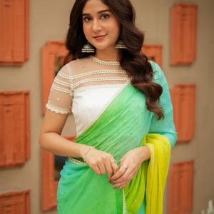 May include: A woman wearing a green and blue saree with a white blouse. The saree has a gradient effect, transitioning from green to blue, with a yellow border. The blouse has a sheer neckline and short sleeves. The woman is wearing silver earrings.
