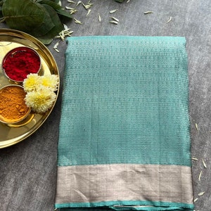 May include: A folded teal green saree featuring a subtle geometric pattern across the fabric and a wide, metallic silver border along the bottom edge. The textile has a textured appearance and is presented in a neat, rectangular stack.