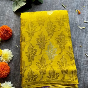 May include: A folded mustard yellow fabric featuring a repeating dark yellow fern leaf pattern. The textile has a subtle sheen and a smooth texture, with a visible horizontal fold line near the bottom edge and a hint of purple fabric tucked inside.
