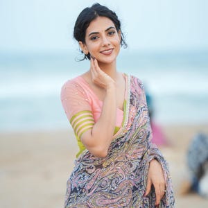 May include: A woman wearing a pink blouse with sheer sleeves and a paisley patterned saree. The saree features a mix of pink, black, and yellow colors. The background shows a beach and the ocean.