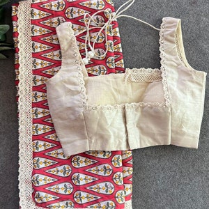 May include: A cream-colored blouse with a square neckline and lace trim, paired with a red and white patterned fabric with a lace border. The fabric features a repeating floral design.