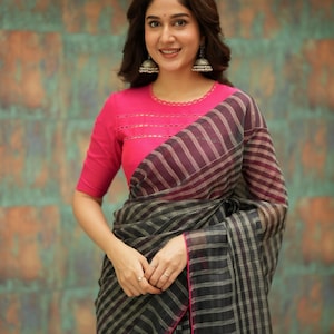 May include: A woman wearing a pink blouse with a striped saree. The saree is primarily black and gray with pink trim. The blouse has a round neckline and short sleeves. The woman is also wearing silver earrings.