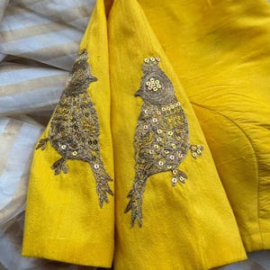 May include: A yellow blouse with embroidered bird designs on the sleeves. The birds are outlined in gold thread and have sequins for accents.