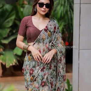 May include: A saree featuring a light grey base with a vibrant floral print in shades of red, yellow, and green. The garment is paired with a short-sleeved, V-neck blouse in a solid dark maroon color. The text THREADSLABEL is visible on the fabric.