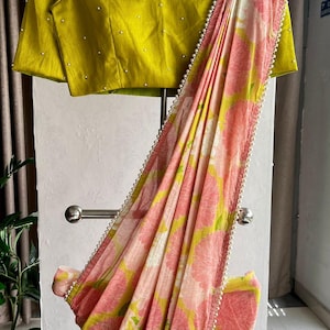 May include: A pink and green floral saree with a pearl trim. The saree is draped over a hanger and has a matching green blouse with pearl embellishments.