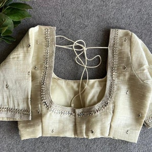 May include: A cream-colored blouse with short sleeves, featuring a square neckline adorned with silver metallic embroidery and small reflective mirror accents. The garment includes thin fabric ties at the back and scattered mirror embellishments.