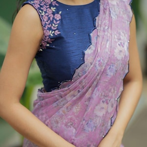 May include: A sleeveless navy blue top with floral embroidery on the shoulder and a scalloped neckline. A sheer, floral-patterned lavender and pink fabric drapes over the top. The fabric has a scalloped edge.