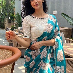 May include: A teal saree with a floral pattern and a white blouse. The saree features a large floral print in shades of white and beige. The blouse is white with mirror embellishments around the neckline and sleeves. The saree is draped over the body.
