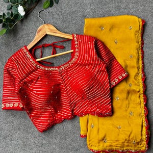 May include: A red and gold embroidered blouse with short sleeves and a matching yellow saree with a red border. The saree has a scalloped edge and is decorated with gold embroidery.