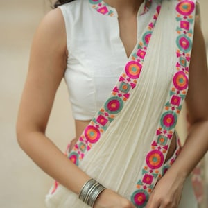 May include: White sleeveless top and off-white saree with a colorful border. The saree features a decorative border with pink, orange, and teal geometric patterns. A silver cuff bracelet is worn on the wrist.